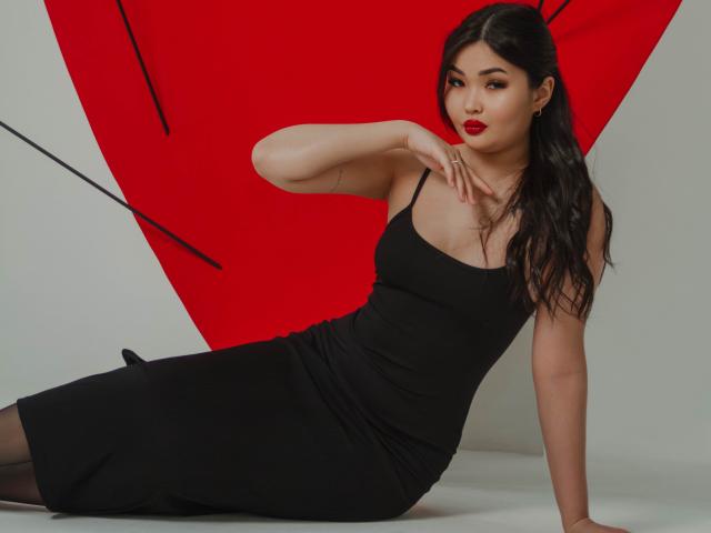 TeyaFaber has dark hair, red lipstick, and wears a black dress, sitting with one hand near her face, showing off her tits and ass in a sexy pose.