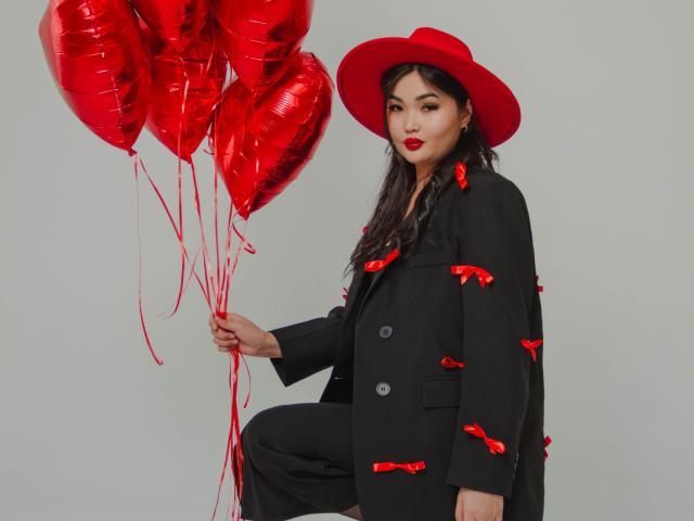 TeyaFaber with red heart balloons, black coat, red ribbons, and red hat, holding balloons, long black hair, red lipstick, standing, gray background, looking at camera.