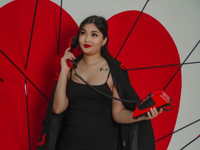 TeyaFaber with red lipstick, black dress, holding red and black phone, black jacket, tattoos on chest, standing in front of large red heart with black lines.