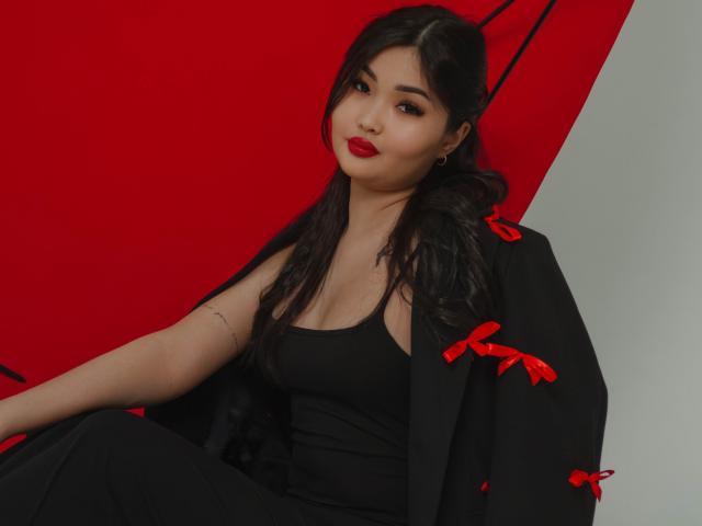 TeyaFaber has dark hair, red lipstick, and is wearing a black dress with a black shawl that has red ribbons. She has a tattoo on her left arm.
