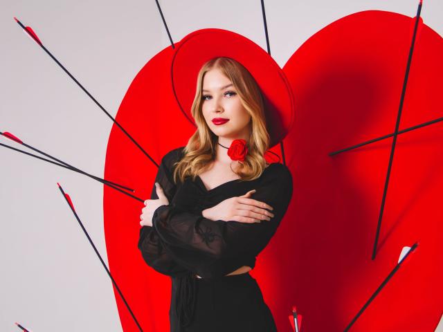 NicoleBrian, avec ses cheveux blonds, son chapeau rouge, sa robe noire et son collier de roses, les bras croisés, affichant une attitude confiante, entourée d'accessoires en forme de cœur rouge et de flèche noire. Ses seins sont partiellement visibles sous la robe..