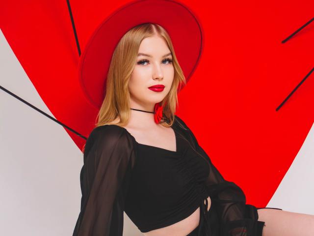 NicoleBrian con capelli biondi, cappello rosso, top nero trasparente, rossetto rosso, seduta, che mostra il seno, collana nera, sfondo bianco, motivo triangolare rosso dietro di lei..