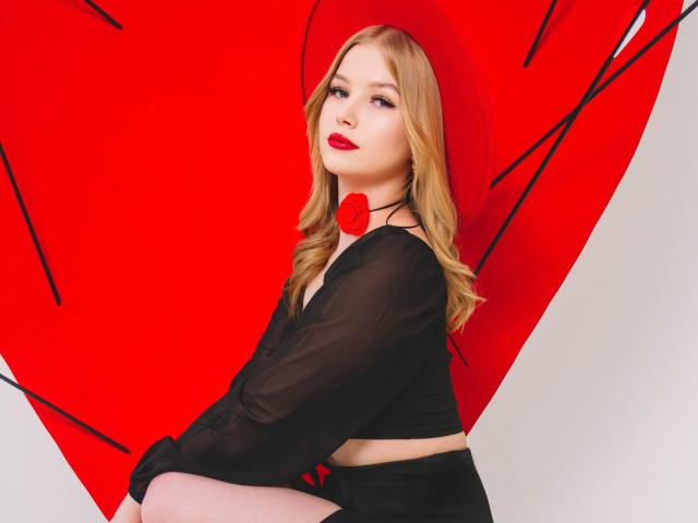 NicoleBrian met blond haar, rode lippenstift, een zwarte doorschijnende top en een rode rozenketting, zittend tegen een grote rode hart, kijkend naar de camera met een subtiele glimlach..