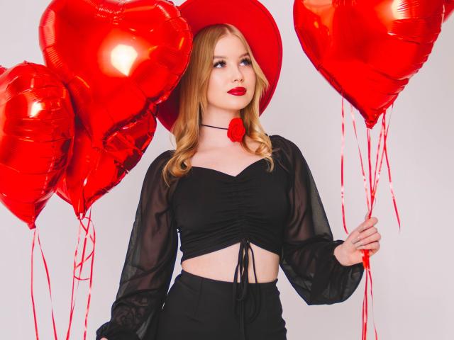 NicoleBrian con palloncini a forma di cuore rossi, indossa una maglia nera trasparente, una gonna nera e un cappello rosso, tiene in mano due palloncini, ha il rossetto rosso, i capelli biondi e una collana con una rosa rossa..
