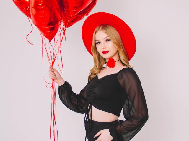 NicoleBrian with blonde hair, red hat, black sheer top, holding red balloons, showing a cute face with red lipstick, and a small red bow on her neck.