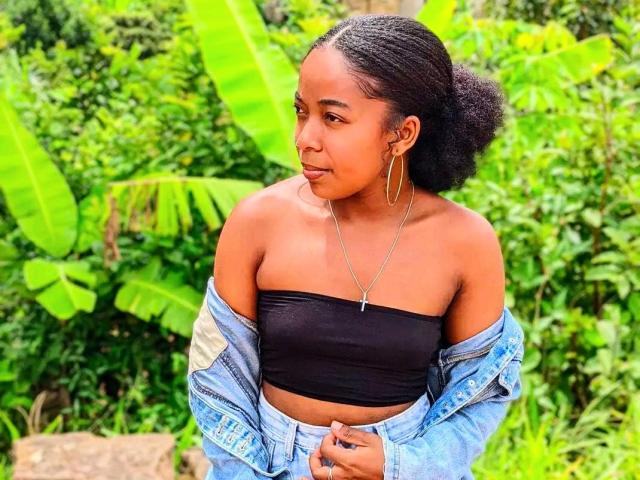 Edminee with dark skin and curly hair, wearing a black tube top, blue denim jacket, and gold hoop earrings, looking to the side, standing in greenery.