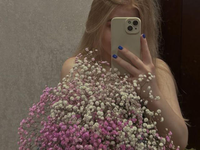 Kisa69Hot with blonde hair, blue nails, holding pink and white baby's breath flowers, taking a mirror selfie with an iPhone, covering her tits.