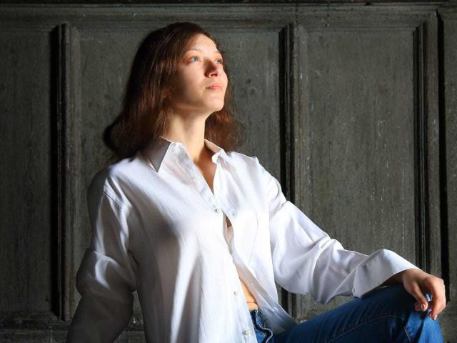 Silerta, con cabello castaño, una camisa blanca abotonada y jeans azules, sentada contra una pared oscura, mirando hacia arriba, con la luz del sol en su rostro, la mano derecha sobre la rodilla, en una pose relajada..