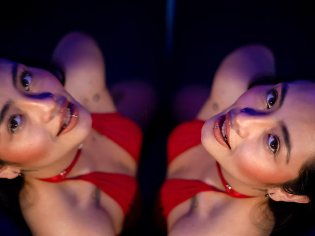 MarieBonnet has reflective, mirrored face showing her red bra, pierced nose, and subtle smile. She's looking at the camera with her green eyes and a slight open-mouth.