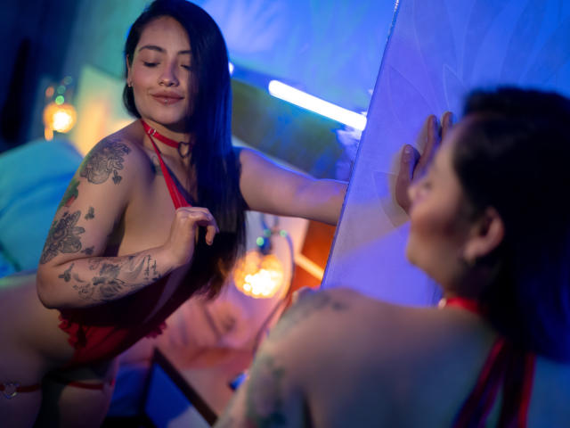 MarieBonnet with tattoos, red lingerie, and a choker, leaning forward, adjusting her strap, looking in a mirror, showing her tits and ass, in a blue-lit room.