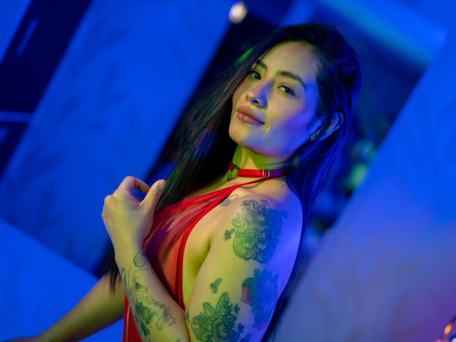 MarieBonnet with long black hair, red sheer dress, tattoos on arm, looking over shoulder, smiling, blue and green lighting, holding dress, showing small tits and puffy ass.