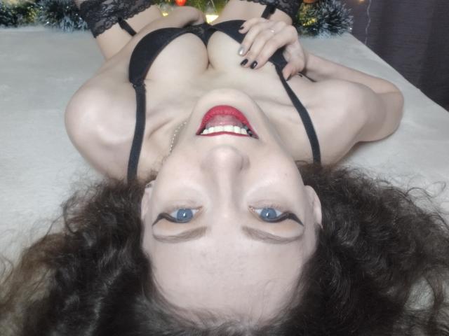 Silerta lies on her back with curly brown hair, blue eyes, red lipstick, black bra, and lace stockings, smiling at the camera. Her tits are prominent, and she's wearing black nails.