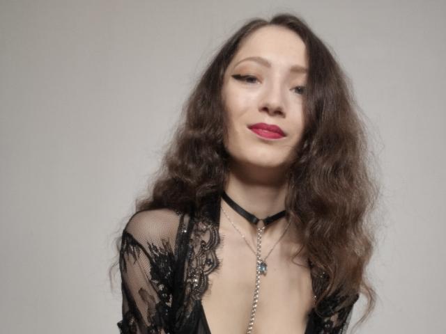 Silerta with wavy brown hair, red lipstick, black lace robe, and choker, showing her tits slightly, wearing a chain necklace with a pendant.