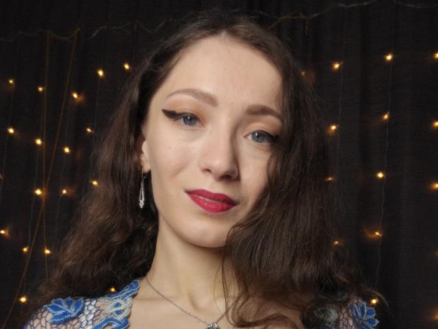 Silerta has wavy brown hair, blue eyes, and red lipstick, with bold black eyeliner. She's wearing a blue patterned top and silver earrings, against a dark backdrop with tiny lights.