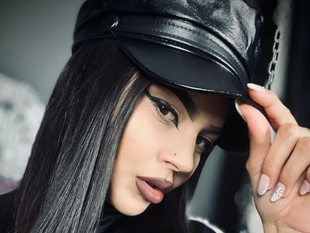 Mistress Edith Fuchs, con lunghi capelli neri, indossa un berretto di pelle nera, un eyeliner nero spesso e un rossetto nude, e tocca il berretto con una mano che mostra un anello scintillante..
