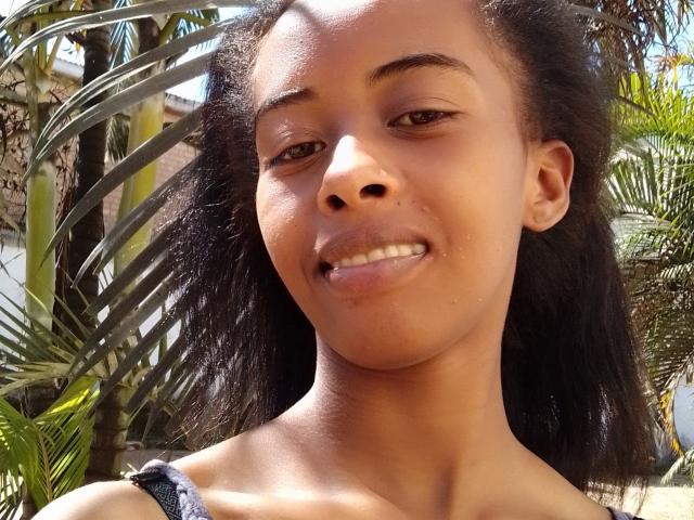 AmourSexy69's face with dark brown skin, small nose, and slight smile, showing her teeth. She has curly black hair and a slight tan, standing in front of green plants.