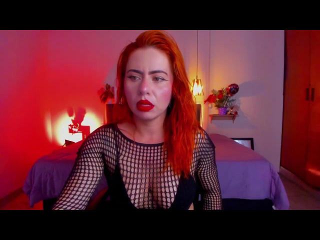 DulceCharlotteHot with red hair, wearing a black fishnet top, showing her tits, red lipstick, and a serious expression in a room with red and yellow lighting.