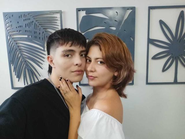 JamesAndRouse, with short brown hair, black shirt, and chain, has a hand on JamesAndRouse's white off-shoulder top, brown hair, and black nails. Three blue leafy wall pieces.