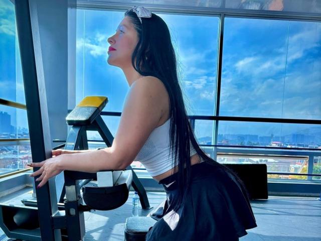 KinkyVane with long black hair, white crop top, black skirt, and checkered headband, kneeling on gym machine, showing her ass, looking to the side, in bright sunlight.