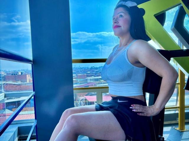 KinkyVane with big tits, wearing a gray sports bra and black Nike shorts, sitting on an exercise machine, looking up, with blue sky and clouds outside.
