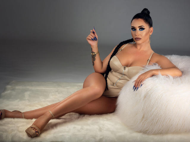 MissElenya, con grandes senos, piel morena, ojos azules, cabello negro recogido en un moño, vestida con un vestido beige, sosteniendo una uña, sentada en una alfombra blanca y esponjosa, con las piernas cruzadas, y usando zapatos de tacón beige..