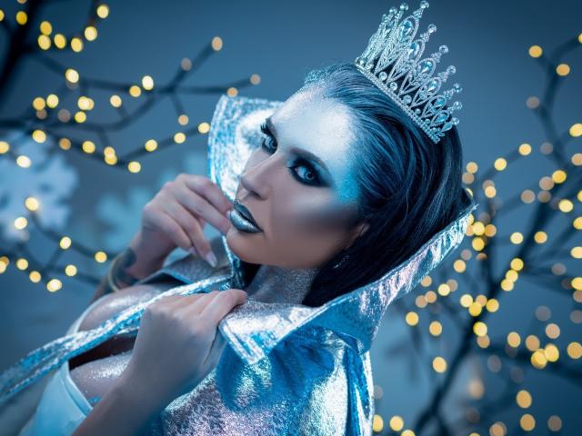 MissElenya, avec son maquillage argenté, son cape bleu métallique et sa couronne argentée scintillante, tenant son collier. Son rouge à lèvres noir et son maquillage bleu des yeux ressortent. Elle porte une tiare argentée..