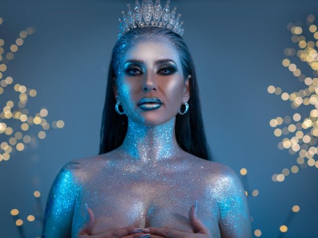 MissElenya, avec son visage et son corps scintillants de bleu, portant une couronne étincelante, cachant ses seins avec ses mains, avec des boucles d'oreilles lumineuses, des cheveux foncés et un maquillage intense, sur un fond bleuâtre..