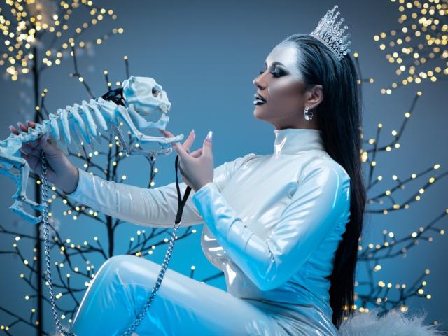 MissElenya with silver crown, white latex dress, and earrings, holding a skeleton skull with a chain, smiling, black hair, blue-gray background, and twinkling lights.