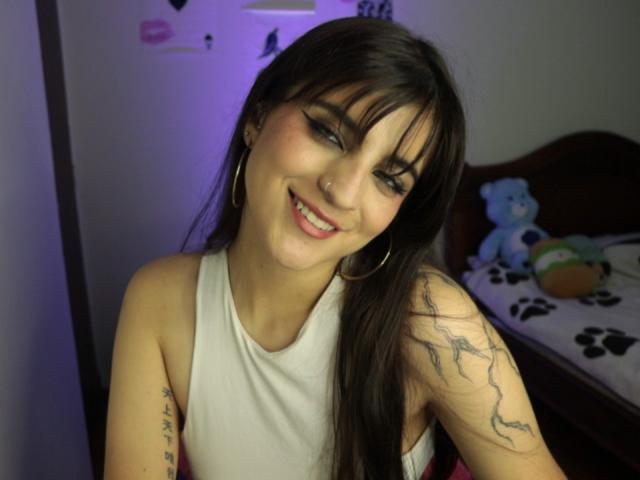 SarahhTanyas with dark brown hair, bangs, and hoop earrings, smiling, wearing a white tank top, showing a tattoo on her right arm, and sitting on a bed.