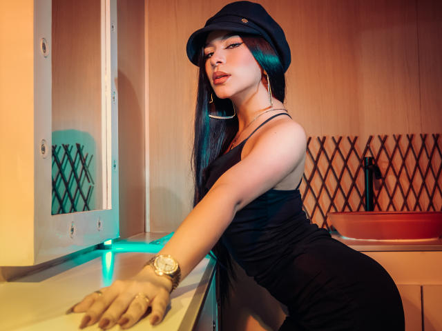ZoeCarlter with long black hair, wearing a black dress, cap, and gold watch, leaning on a counter, showing her ass, with a confident, sexy expression.