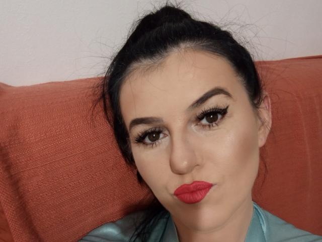 HazelR has dark hair in a ponytail, wearing red lipstick, with thick black eyebrows and long eyelashes, sitting against a red pillow, looking at the camera.