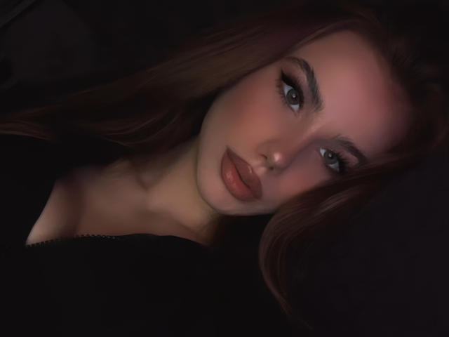 NicoleNight has pouty pink lips, green eyes, and long brown hair. She's looking at the camera with a soft, sexy expression, wearing a black top.