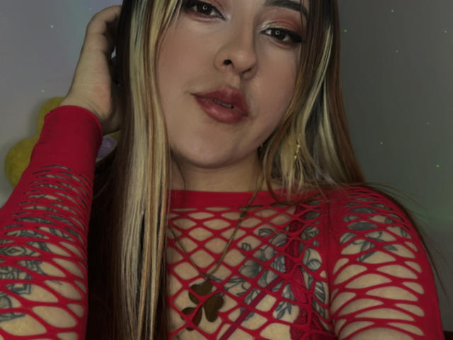 SarahRusso with blonde and brown hair, wearing a red fishnet top, exposing her tits and tattoos, has her hand in her hair, and is looking at the camera.