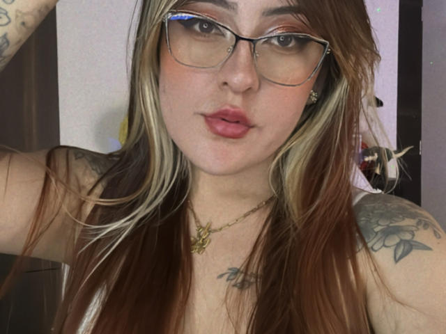SarahRusso with glasses, pouty lips, and brown/ blonde hair, tattoos on arms, wearing a gold necklace, looking at camera. Boobs partially visible, topless.