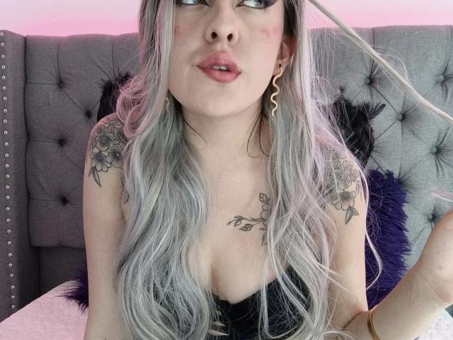 SarahRusso with long gray hair, tattoos on shoulders and chest, wearing black top, looking up, pinkish light in background, purple and gray headboard, wavy hair, pouty lips.