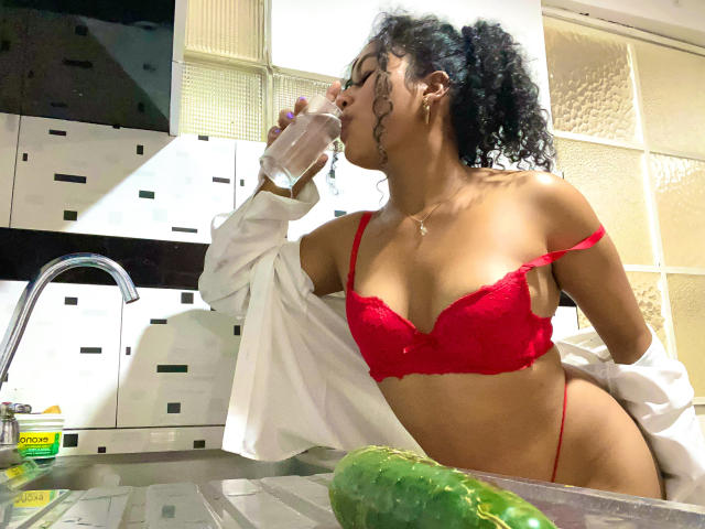 SophieSonyas with curly black hair, drinking water, wearing red lace bra and thong, white shirt off, in kitchen, green cucumber on counter, red bra highlights her small tits.