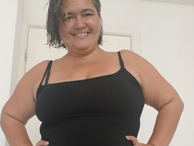 Bethina69 with wet brown hair, smiling, wearing a black spaghetti-strap top, hands on hips, showing her tits, standing in a white room with a door in the background.