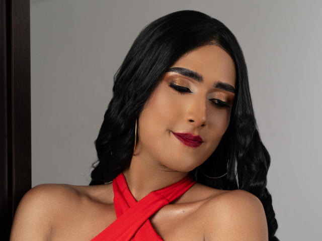 FallonRogers with black wavy hair, wearing a red halter top, gold hoop earrings, and red lipstick, eyes closed, smiling slightly, soft brown skin, looking relaxed.