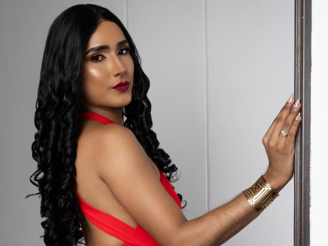 FallonRogers with long black curly hair, red halter top, gold bracelet, and ring, looking over shoulder, touching wall, red lipstick, tan skin, sexy, attractive.