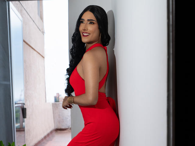 FallonRogers with long black curly hair, red dress, and smiling, showing her ass and tits, leaning against a white wall, looking over her shoulder, red lipstick, cute.