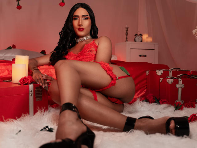 FallonRogers in red lace lingerie, sitting on a white furry rug, legs crossed, showing her ass, black high heels, long black hair, red lipstick, and a silver choker.