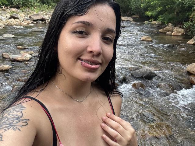 SarahGood with wet black hair, tongue out, small tattoo on shoulder, holding chest, wearing red-black strap top, standing by rocky stream. She's smiling, water droplets on face.