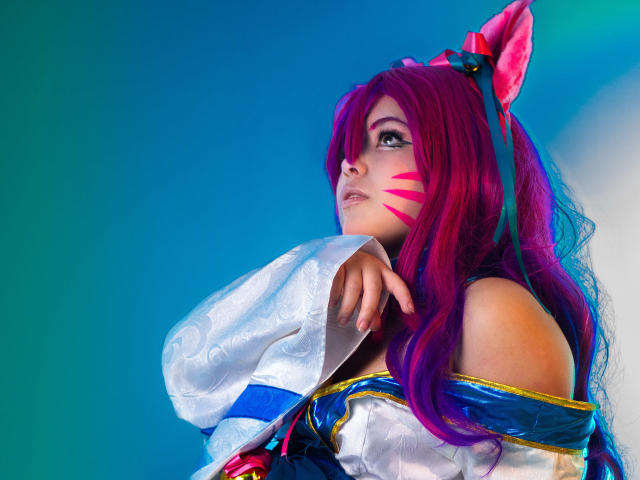 HoneyBonny with bright pink and purple hair, cat ears, white and blue outfit, pink facial stripes, looking up, right hand near chin, blue and green gradient background.