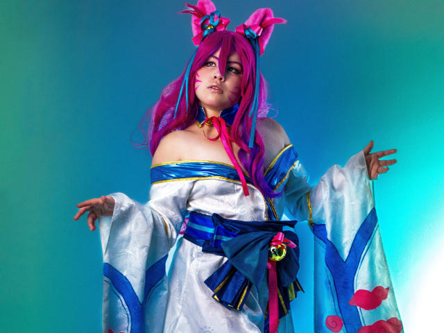 HoneeyBonny in a pink-haired cosplay with rabbit ears, white off-shoulder kimono, blue ribbon, and pink heart patterns, holding out arms, looking up with a cute, confident expression.