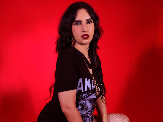 DianaClarke has dark wavy hair, red lipstick, and wears a black shirt with a graphic, showing her tits slightly, against a bright red background. She's sitting with fishnet legs.