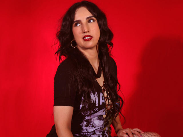 DianaClarke with long black wavy hair, red lipstick, black top, and hoop earrings, looking to the side with a slight smile, against a bright red background.