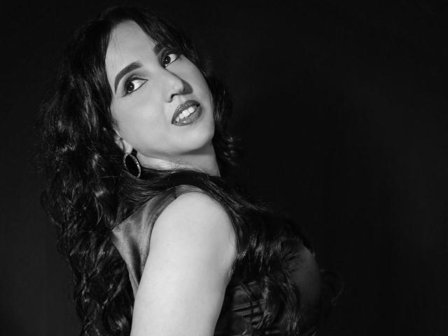 Black-and-white photo of DianaClarke with long, wavy dark hair, looking over her shoulder, wearing a sleeveless top, hoop earrings, and glossy lipstick, showing her ass slightly.