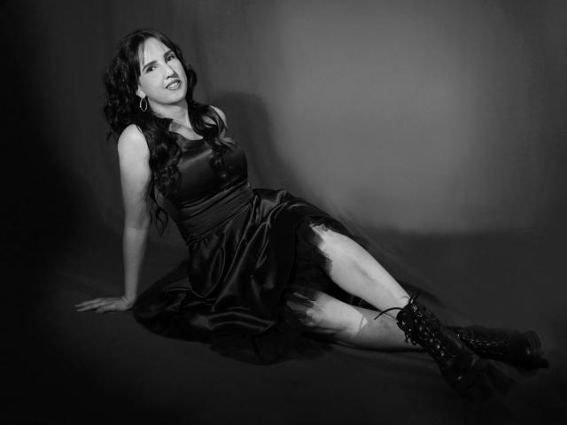 DianaClarke is sitting with her back against the floor, wearing a black satin dress and boots, her legs spread, looking up with a slightly annoyed expression.