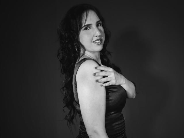DianaClarke with curly black hair, smiling, wearing a black leather dress, covering her small tits with one hand, dark gray background, black and white photo.