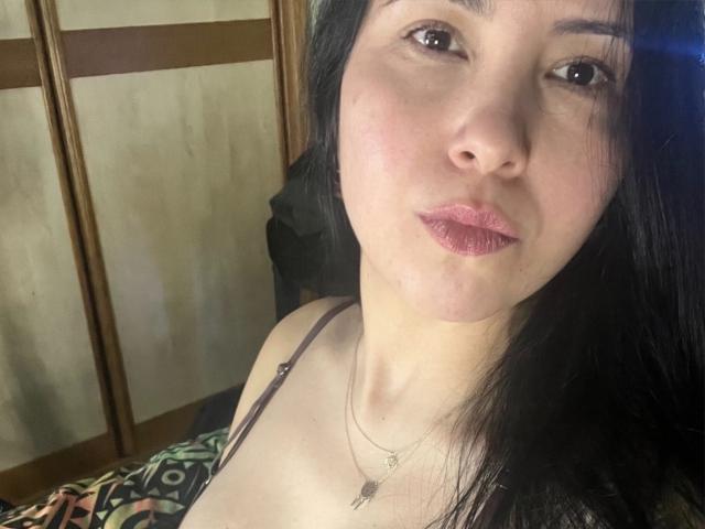 ValeriaQ has pale skin, black hair, brown eyes, and pink lipstick. She's wearing a spaghetti strap top with a necklace, and her tits are slightly visible. She's pouting.